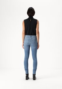 Top preto sem mangas com detalhe em forma de chave nas costas, combinado com jeans justos azuis claros e botas de cano curto pretas, exibindo uma silhueta elegante.