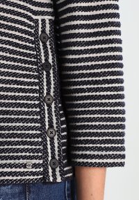 Cardigan rayé bleu marine et blanc avec un tissu texturé, doté de boutons foncés et d'un design à manches trois-quarts.