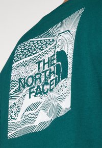 Turfarget bomulls t-skjorte med et hvitt grafisk design med bølgende linjer, teksturer og teksten "THE NORTH FACE". Rund hals, korte ermer.