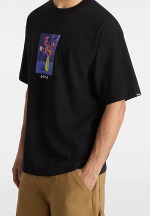 Mies, jolla on päällään musta Vans-t-paita, jossa värikäs abstrakti kukkakuosi, ja vaaleanruskeat housut, käsi taskussa, vartalo näkyvissä, vaalea tausta.