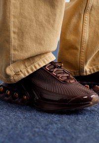 Brown athletic shoes featuring a mesh upper, rounded toe, and distinctive patterned design on the sole with rounded accents.