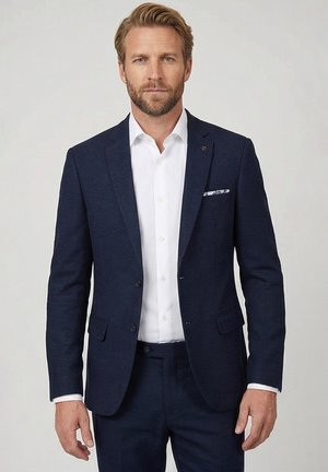 Blonde man met baard, gekleed in een marineblauw pak, wit overhemd en pochet, staande tegen een effen lichte achtergrond.