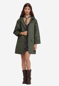 Grøn, knælang jakke med hætte, knaplukning, to frontlommer og oprullede ærmer. Båret over en blomsterkjole og brune støvler.