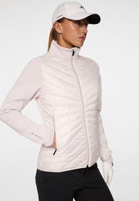 En kvinna med långt hår bär en ljusrosa zip-jacka och en vit keps, poserar självförtroende med en hand på höften framför en enkel bakgrund.