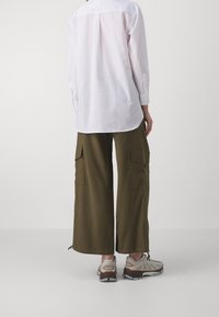 Persona che indossa una camicia bianca a maniche lunghe, pantaloni cargo larghi color oliva con tasche e sneakers beige, in piedi contro uno sfondo semplice.