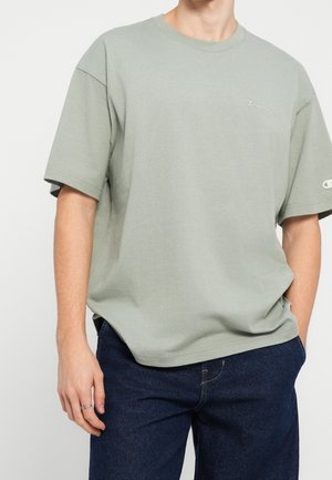 Osoba nosząca jasnuzielony, oversize t-shirt Champion z logo na piersi i rękawie, w zestawieniu z ciemnoniebieskimi jeansami, stojąca z jedną ręką w kieszeni.