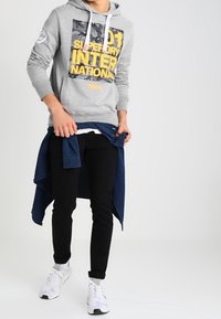 Hombre con sudadera con capucha gris y texto amarillo, pantalones negros, zapatillas blancas y una camisa azul atada a la cintura.