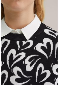 Juodas megztinis su balta širdies raštu, briaunuotu apykakle ir po juo baltais marškiniais su apykakle. Audinys atrodo megztas ir minkštas.