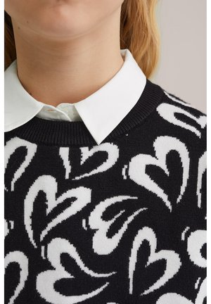 Pull noir avec un motif cœur blanc, col côtelé, et chemise blanche à col en dessous. La texture semble tricotée et douce.