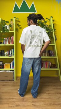 Balta grafiska T-krekls ar kaķa dizainu un tekstu "MATCHA The Puma Way," kopā ar vaļīgām zilām džinsa biksēm.