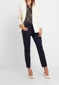 Blazer doppiopetto avorio sopra una blusa blu navy a fantasia e pantaloni slim-fit blu navy. Un paio di scarpe con tacco alto bianche completano l'insieme.