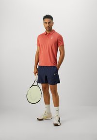 Short-sleeve pink polo, navy shorts, white socks, and multi-colored tennis shoes. Holding a tennis racket. Simple, modern athletic design.