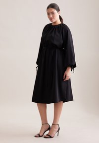 Czarna sukienka midi z marszczonym dekoltem, długimi rękawami i paskiem w talii. Miękki materiał o gładkiej fakturze. W zestawieniu z czarnymi szpilkami.