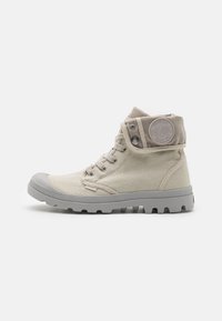 Beige canvas high-top shoes with gray rubber toe cap and sole, featuring a round logo patch and tonal laces. Textured upper with padded collar.
