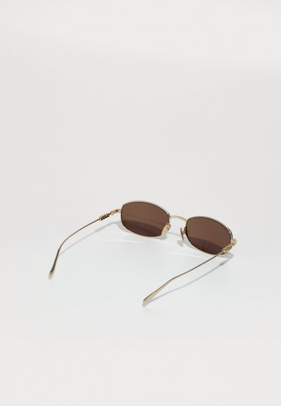 Gafas de sol ovaladas con lentes marrones, montura de metal dorado, accesorios decorativos en las patillas y brazos delgados y curvados.