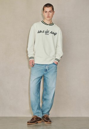 Young man wearing a white sweatshirt with black text, light blue jeans, brown shoes, standing against a plain beige wall with one hand in pocket.