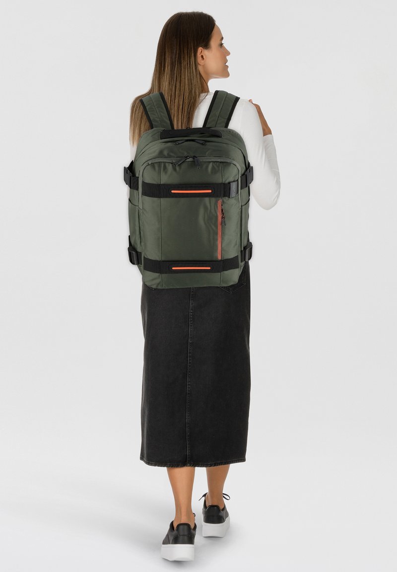 Mochila de tela verde con correas dobles, acentos negros, múltiples compartimentos con cremallera y un destacado detalle de cremallera roja.