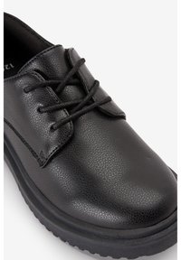Chaussure noire à lacets en matériau synthétique texturé, avec un bout rond et une semelle en caoutchouc épaisse pour un confort accru.