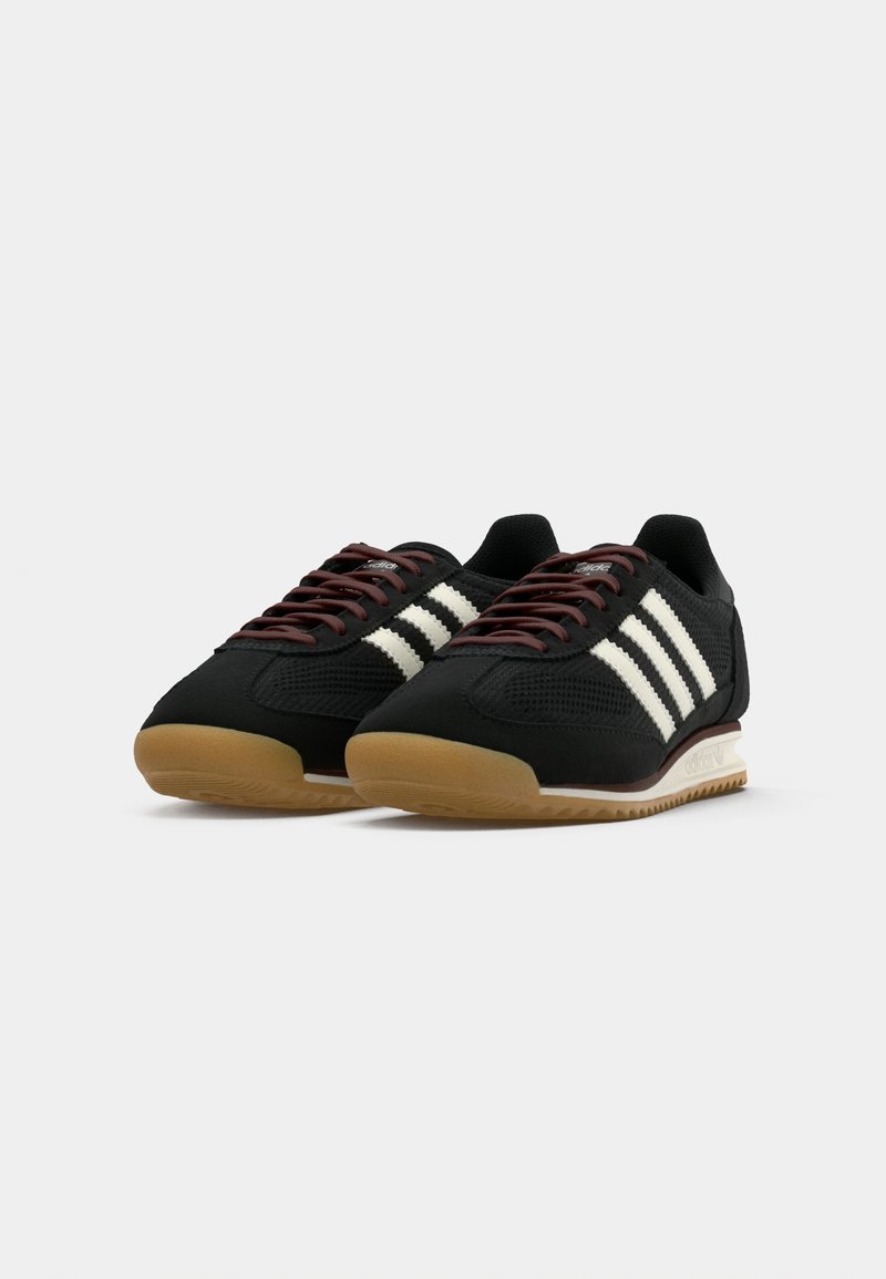 Zapatillas deportivas negras con rayas blancas, parte superior de malla texturizada, cordones marrones y suela de goma con un patrón de zigzag.