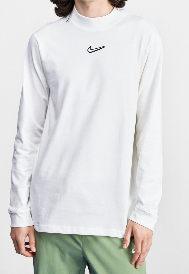 Camiseta de manga larga blanca hecha de algodón. Presenta un cuello alto y un logo de Nike negro bordado en el pecho. Diseño simple y textura suave.