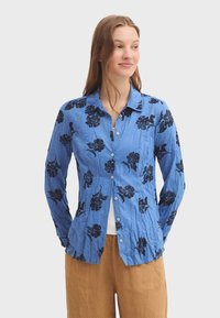 Chemise bleue à boutons en tissu léger, ornée de motifs floraux noirs, manches longues et col. Texture froissée.
