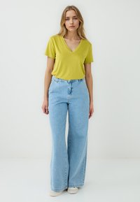 Una modella indossa una brillante maglietta gialla con scollo a V e jeans larghi di un azzurro chiaro, con i capelli lunghi che incorniciano il suo viso su uno sfondo bianco semplice.