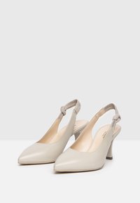 Tacchi slingback in pelle beige con punta affusolata, un'accessorio annodato sulla cinghia e un tacco quadrato di media altezza. Texture liscia con una finitura lucida.