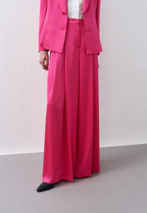 Άτομο που φοράει φωτεινό ροζ παντελόνι σε ευρεία γραμμή και αντίστοιχο σακάκι πάνω από λευκό πουκάμισο, στέκεται με το ένα χέρι ορατό.