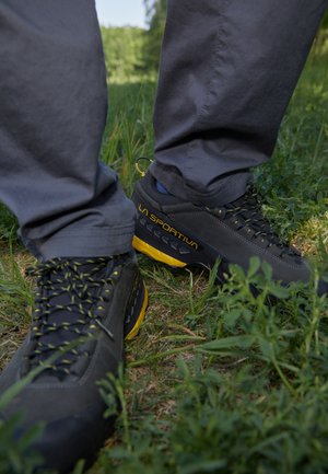 Scarpe da trekking nere e grigie con accenti gialli, dotate di una suola in gomma testurizzata, punta rinforzata e lacci decorati, posizionate sull'erba.