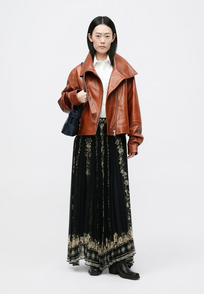 Smeđa kožna jakna s širokim okovratnikom, bijela košulja i crna cvjetna maksi suknja. Model drži malu crnu torbicu; nosi crne gležnjače.