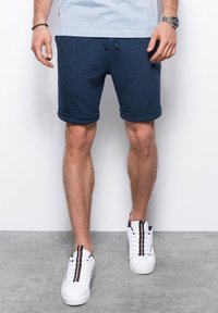 Tmavě modré látkové šortky s ohrnutými manžetami, šňůrkovým páskem a texturovaným designem, spárované s bílými teniskami s barevnými pruhy.