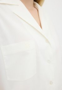 White button-up shirt with a smooth texture, notched collar, and a left chest pocket featuring subtle embroidered detailing.