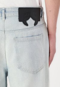 Des jeans en denim bleu clair présentent une patch en cuir noir à l'arrière avec un design distinct, mettant en valeur des coutures uniques et une surface texturée.