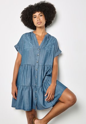 Kvinde med krøllet afro-hår iført en blå denimkjole med knapper foran og broderede skuldre, poserer med det ene knæ løftet mod en hvid væg.