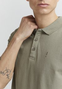 Olivengrønn polo med tre knapper, korte ermer, teksturert stoff og en liten logo på brystet.