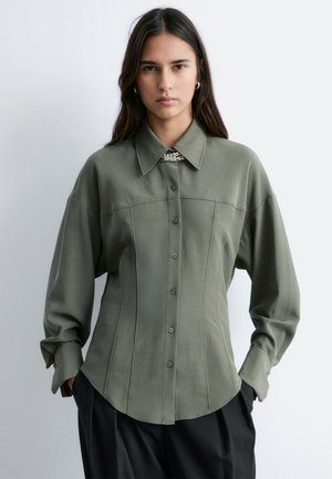 Młoda kobieta o długich ciemnych włosach ubrana w dużą oliwkowo-zieloną koszulę na guziki z szerokim kołnierzem oraz czarne spodnie, stojąca na białym tle.