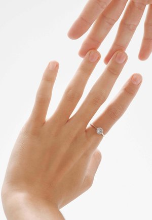 Anillo de plata con una gema clara y circular, mostrado en una mano con uñas naturales sobre un fondo claro. La mano se extiende hacia otra mano.