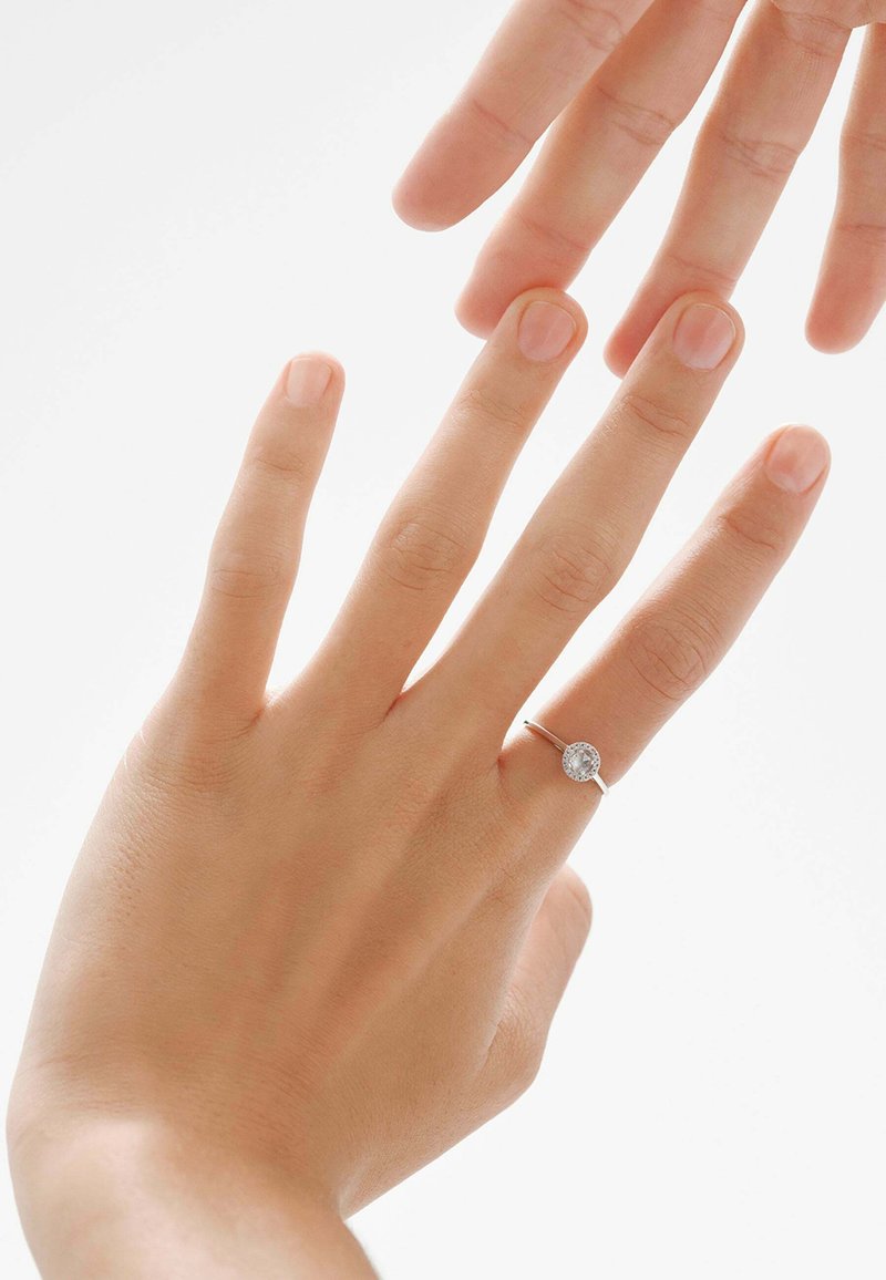 Anillo de plata con una gema clara y circular, mostrado en una mano con uñas naturales sobre un fondo claro. La mano se extiende hacia otra mano.