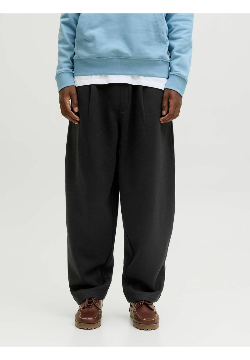 Pantalones de chándal anchos y negros, hechos de una tela suave, con cintura alta y pliegues sutiles, combinados con zapatos marrones de cordones y una sudadera azul.