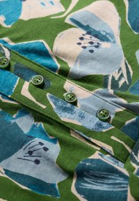 Tejido de algodón floral en tonos verdes y azules con acentos en color crema, que presenta botones de plástico verdes a lo largo de un tapete frontal.