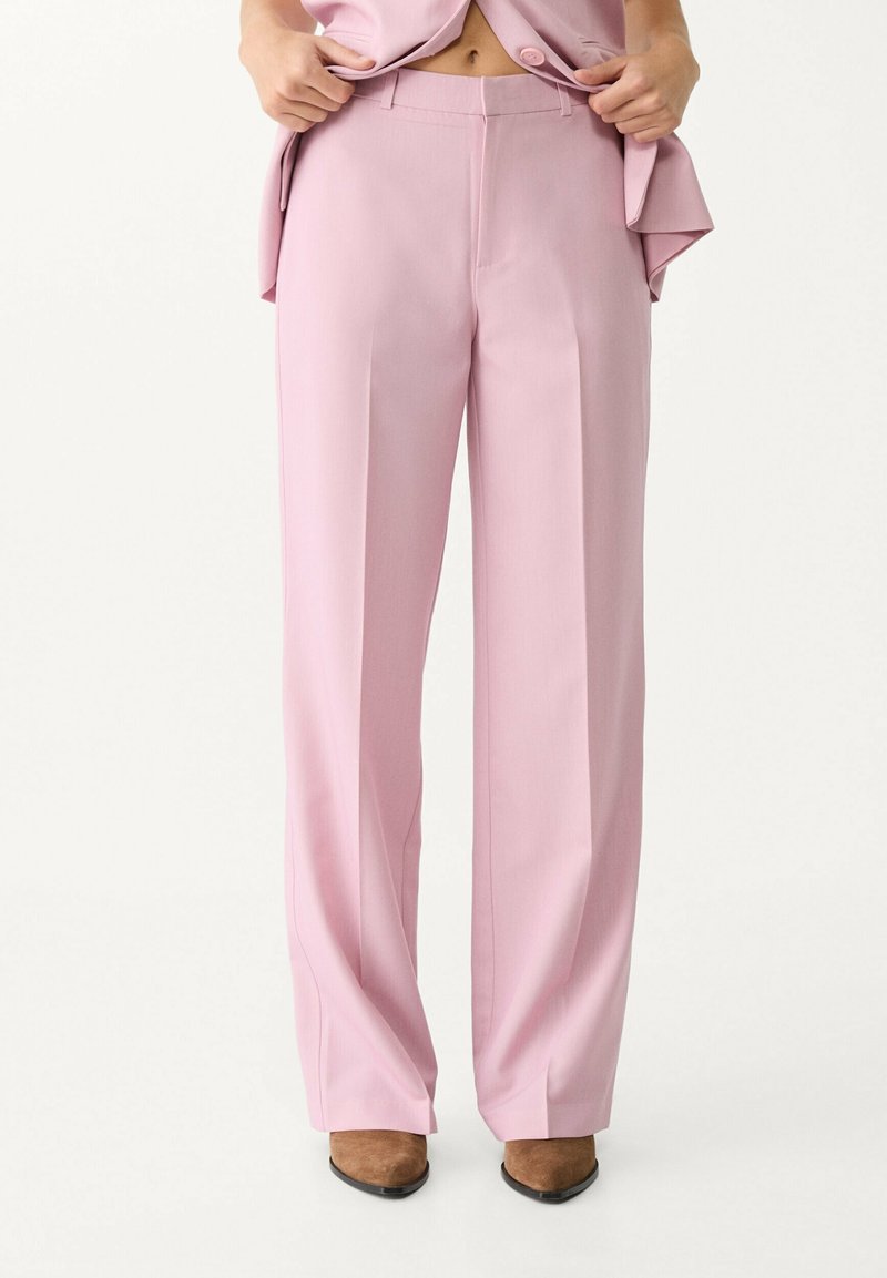 Personne portant un pantalon large taille haute rose clair avec des chaussures brunes pointues, ajustant un blazer rose assorti.
