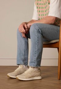 Beige Wildlederschuhe mit weißen Sohlen, die eine glatte Textur und ein minimalistisches Design aufweisen. Getragen mit hellblauen Jeans, sitzend auf einem Stuhl.