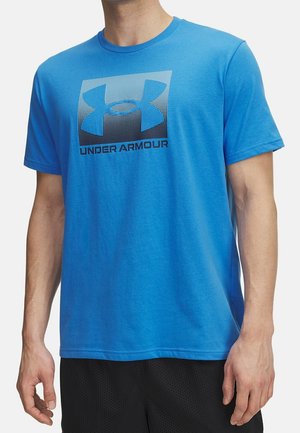 Mężczyzna ubrany w niebieską koszulkę Under Armour z krótkim rękawem, z czarnym logo i napisem na piersi, w połączeniu z czarnymi spodenkami.