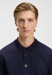 Navy polo shirt with a structured collar, three-button placket, and a rectangular black logo tag that reads "HUGO." Smooth texture, casual fit.