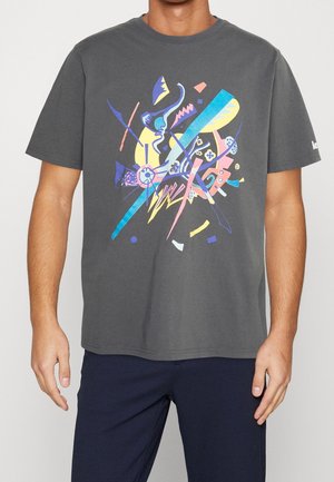 Mand iført mørkegrå T-shirt med farverige abstrakte geometriske former på fronten, kombineret med mørkeblå bukser.