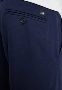 Pantalon bleu marine en tissu texturé, doté d'une poche arrière avec un bouton noir et d'une petite étiquette de marque au-dessus.