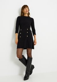 Robe noire côtelée avec un corsage ajusté, une jupe en ligne A et des accents de boutons dorés. Le modèle porte des bottes en cuir noir montant jusqu'au genou.