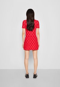 Abito rosso in maglia con maniche corte, motivo a diamante e corpino aderente, abbinato a scarpe nere. Sfondo minimalista.