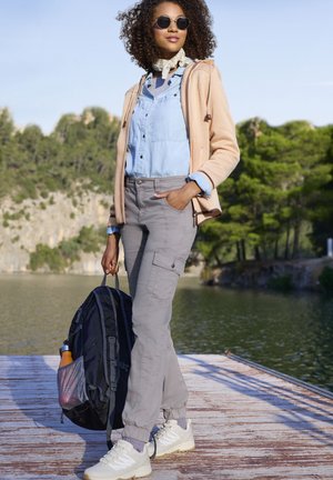 Heller Pfirsichfarbener Zip-Up-Hoodie über einem blauen Hemd, graue Cargo-Hosen, weiße Sneakers und hält einen schwarzen Rucksack am See.