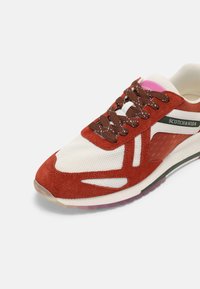 Baskets blanches et rouge rouille avec des lacets marron, des matériaux en mesh et en daim, et un logo Scotch & Soda sur le côté.